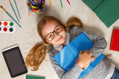 Cute little girl in glasses with a book lying on the floor. A child is surrounded by a book, tablet, paints, brushes, pencils.